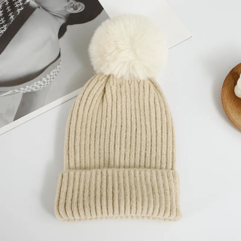 Berretto beige con risvolto a coste e grande pompon bianco panna, fotografato su una superficie chiara