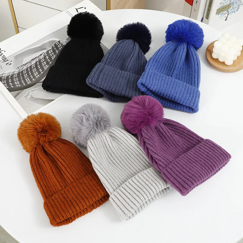 Sei berretti a coste con pompon, neri, blu scuro, blu, ruggine, grigio chiaro e viola, disposti in piano