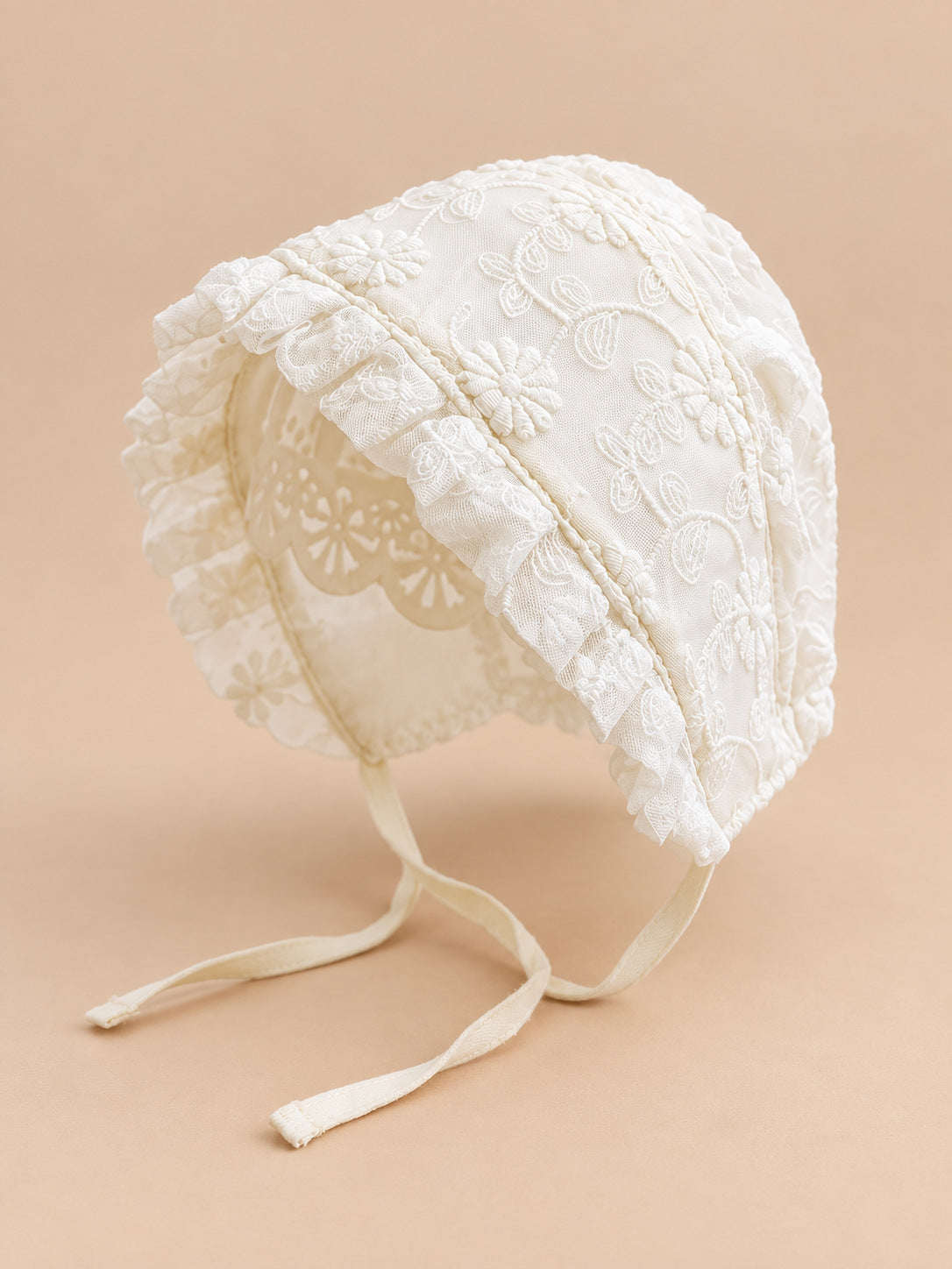 Cuffietta da neonata in pizzo ricamato, modello 02, vista di tre quarti, su sfondo color sabbia e pesca